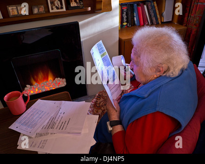 Allocation de chauffage aîné aîné indépendant 90s année dame à la maison devant le feu à l'aide d'une loupe pour lire ses dernières factures d'énergie du ménage Banque D'Images