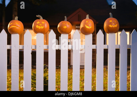 Jack-O-Lantern Visages coincé sur le dessus de l'Picktet blanc clôture avec une maison en arrière-plan au crépuscule au cours de l'automne en Alaska. Banque D'Images