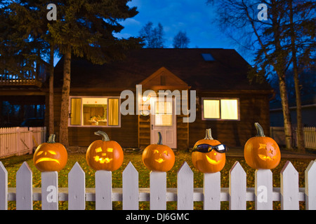 Jack-O-Lantern Visages coincé sur le dessus de l'Picktet blanc clôture avec une maison en arrière-plan au crépuscule au cours de l'automne en Alaska. Banque D'Images