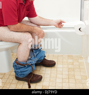 Homme assis dans la salle de bains sur les toilettes, arrachant un morceau de papier de toilette. Banque D'Images