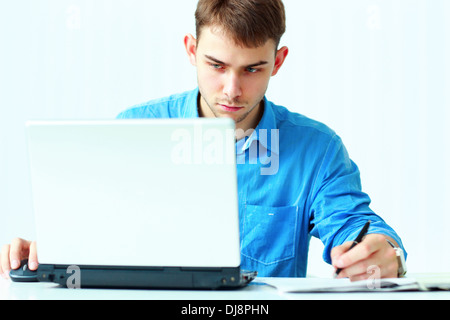 Jeune homme concentré à écrire des notes vers le bas et looking at laptop in office Banque D'Images