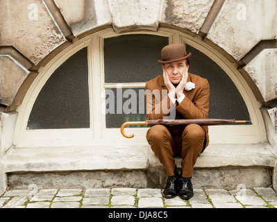 La ville de Gand dans dapper un costume marron et chapeau melon assis par une fenêtre en arc avec sa tête dans ses mains. Banque D'Images