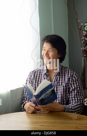 Un jeune homme asiatique assis à une table tenant un livre de voyage souriant et heureux, en regardant par la fenêtre. Banque D'Images