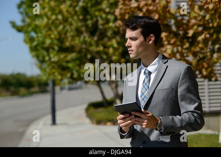 Un jeune homme d'affaires détenant un ipad, tablet, à l'écart de manière ambitieuse. Il est debout sur un sentier en face de quelques arbres. Banque D'Images