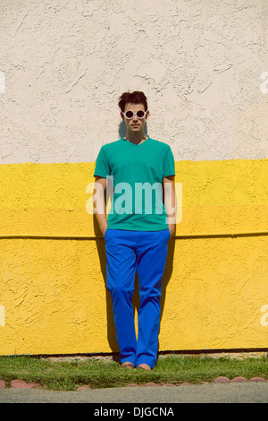 Un jeune homme avec des lunettes ronde,pied contre mur jaune,porte une chemise verte et un pantalon bleu, un concept de mode de couleur Banque D'Images