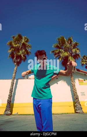 Un jeune homme avec des lunettes rondes et des cheveux funky, permanent, sous ciel bleu entre les palmiers à Los Angeles, Californie Banque D'Images