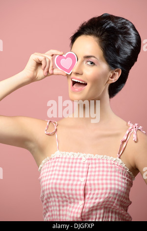 Belle femme, vintage hairstyle tenant un objet en forme de cœur sur son œil et souriant avec enthousiasme. Concept de la Saint-Valentin Banque D'Images