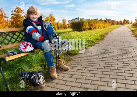 Happy blonde boy putting sur patins à assis sur le banc du parc en automne Banque D'Images