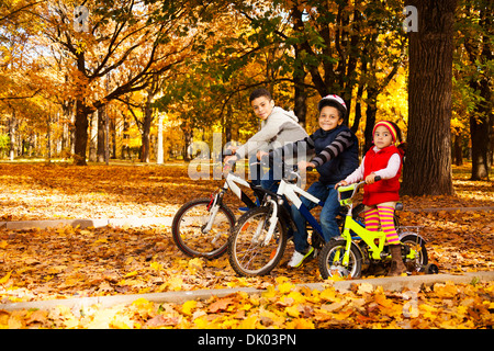 Groupe de trois garçons noirs et les petites filles, frères et soeur la bicyclette et scooter dans l'automne Octobre parc avec feuilles de chêne et d'érable Banque D'Images