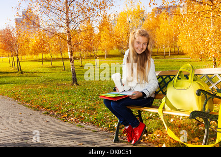 Happy smiling teen blonde girl with long hair holding coffee et les livres et les papiers avec sac portant sur le banc, dans le parc en automne Banque D'Images