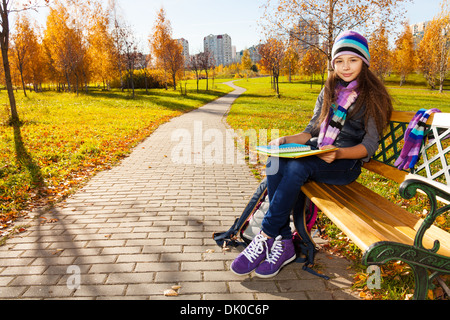 Mignon 11 ans fille dans des vêtements chauds dans le parc assis sur le banc du parc en automne Banque D'Images