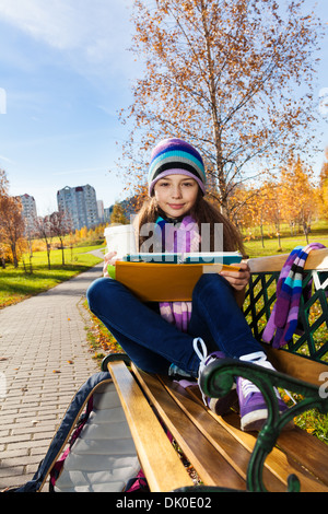 Portrait of smiling nice 11 ans girl holding coffee mug et pour manuels wearing blue purple hat et rhizoctone brun assis sur le banc dans le parc Banque D'Images