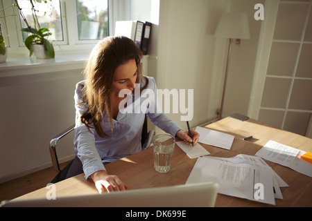Young businesswoman assise à son bureau à écrire des notes à partir d'un ordinateur portable à la maison. Belle femme de race blanche de travail home office Banque D'Images