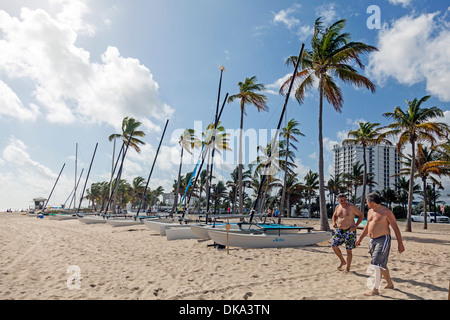 Les hommes de marcher sur la plage de Fort Lauderdale, Floride avec catamaran location de voiliers, de palmiers et de Bahia Mar Hotel au-delà. USA Banque D'Images