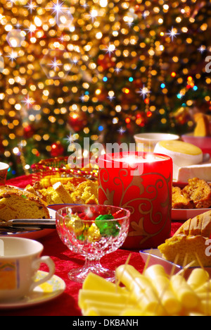 Set de table de Noël traditionnel avec des tasses à thé, des gâteaux, du fromage et une bougie et des lumières scintillantes dans l'arrière-plan. Banque D'Images