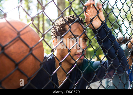 Portrait of young man holding basketball par wire fence Banque D'Images