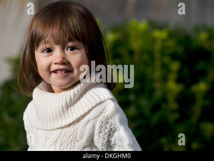 Un enfant de deux ans M. mixed race female sourit tout en ayant sa photo prise. Elle a les cheveux bruns, est vêtu d'un long pull blanc Banque D'Images