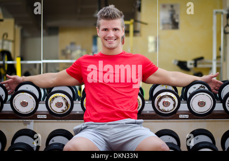 Beau jeune homme dans une salle de sport assis sur rack haltères, souriant et à huis clos. Banque D'Images