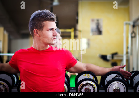 Beau jeune homme dans une salle de sport assis sur les haltères rack, à la recherche d'un côté. Banque D'Images