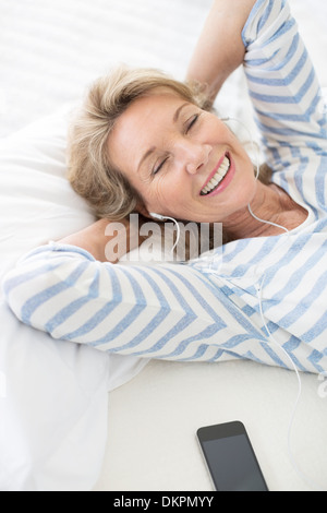 Femme plus âgée listening to earphones on bed Banque D'Images