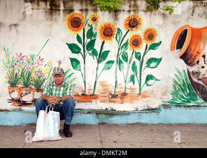 Un homme assis sur la rue par un mur coloré image représentant l'agriculture, La Havane Cuba, Caraïbes Banque D'Images