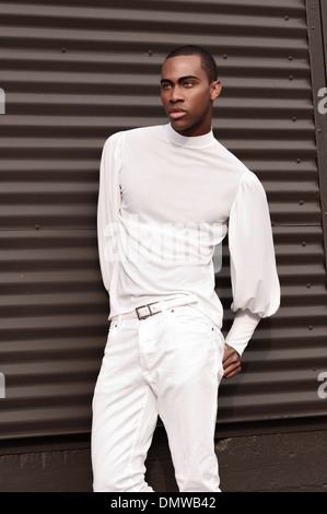 Un bel homme noir, African American male model vêtu d'un costume tout blanc posant contre mur noir métal. Banque D'Images