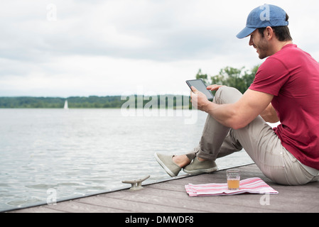 Un homme assis sur une jetée par un lac à l'aide d'une tablette numérique. Banque D'Images