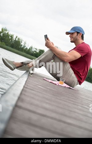 Un homme assis sur une jetée par un lac à l'aide d'une tablette numérique. Banque D'Images