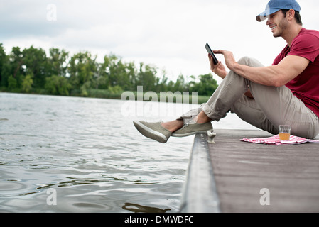 Un homme assis sur une jetée par un lac à l'aide d'une tablette numérique. Banque D'Images