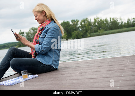 Une femme assise sur une jetée par un lac à l'aide d'une tablette numérique. Banque D'Images