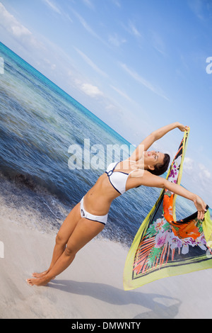 Femme vivace rire dans un bikini au bord de la mer, tenant une écharpe à motifs colorés dans sa main tendue à flottent dans la brise contre une toile de fond de l'océan Banque D'Images