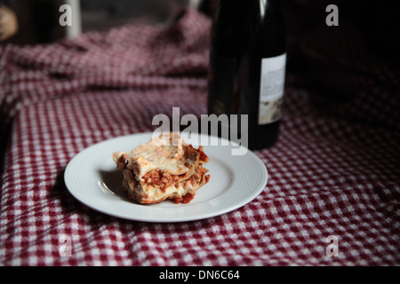 Une tranche de lasagne à la viande fait maison sur une plaque blanche avec une bouteille de vin rouge à l'arrière-plan sur une nappe à carreaux Banque D'Images