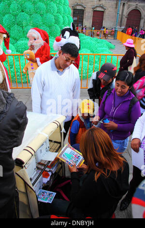 La Paz, Bolivie. 21 décembre 2013. Un assistant du photographe de la rue met une photo des clients qui posent avec le Père Noël et les héros de films populaires dans un cadre en carton. La photo a été imprimée à l'aide d'une imprimante compacte. Dans l'arrière-plan est un arbre de Noël écologique en plastique recyclé des bouteilles de boissons. L'arbre est de plus de 15 m de haut et contient environ 50 000 bouteilles. Credit : James Brunker / Alamy Live News Banque D'Images