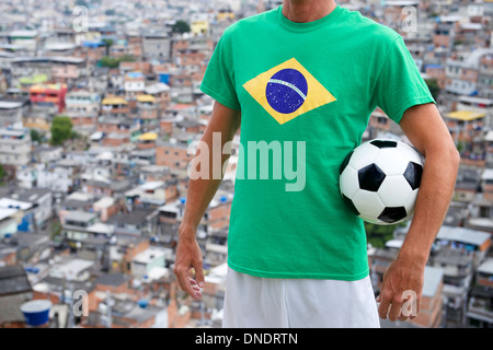 Joueur de football brésilien est en drapeau brésilien t-shirt holding soccer ball en face de l'arrière-plan des bidonvilles favela à Rio de Jane Banque D'Images