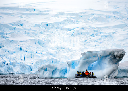 Touristes Zodiac Iceberg Arch Curtis Bay Antarctic Peninsula with People / CURTIS BAY, Antarctique — Expedition touristes dans une manœuvre de bateau gonflable Zodiac à côté d'un arc d'iceberg sculpté dans Curtis Bay, éclipsé par les falaises de glace chaotiques d'un glacier de vêlage face derrière eux. La péninsule Antarctique est l'une des régions qui se réchauffent le plus rapidement sur Terre, et l'accélération du vêlage des glaciers ici a augmenté à la fois le nombre de bergs flottants et le danger imprévisible qu'ils posent pour les petites embarcations opérant à courte distance. Banque D'Images