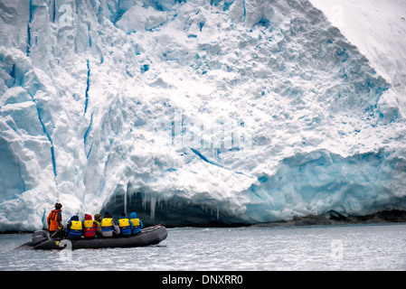 Falaises de glace du glacier touristes du zodiaque Curtis Bay péninsule antarctique avec des gens / CURTIS BAY, Antarctique — Un bateau gonflable du zodiaque transporte les touristes sous les imposantes falaises de glace du glacier sur le rivage de la baie de Curtis sur la péninsule antarctique, des glaçons suspendus à un surplomb d'eau de fonte au-dessus d'eux. Les stries bleues qui traversent verticalement la falaise sont des intrusions d'eau de fonte recongelée - un enregistrement visible de l'eau de surface qui a percolé dans des crevasses et s'est re-gelée, liant les couches de glace annuelles successives et marquant les endroits où le glacier est activement déstabilisé. Banque D'Images