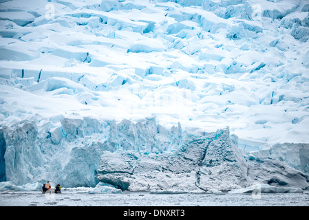 Face de vêlage du glacier de la péninsule antarctique Zodiac Boat Curtis Bay / CURTIS BAY, Antarctique — Un Zodiac transporte des passagers éclipsés par un terminus de glacier fortement crevassé à Curtis Bay sur la péninsule antarctique, avec des blocs de glace tombés vêlés à la ligne de flottaison. La surface chaotique des séracs fracturés au-dessus du front de vêlage marque où le glacier se déplace plus vite que la glace ne peut fléchir, se fissurant sous sa propre contrainte d'écoulement avant de se briser dans la mer. Banque D'Images