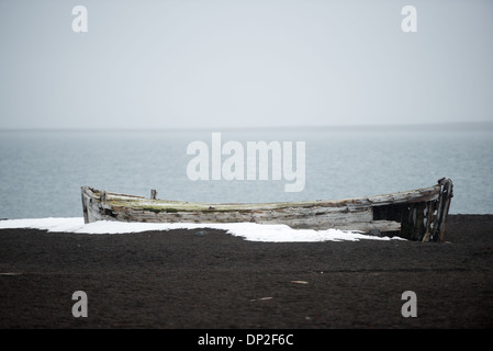 Bateau de pêche abandonné à Whalers Bay, sable noir, île Deception | ÎLE DÉCEPTION, Antarctique — Un bateau à rames en bois désintégré gît échoué sur le sable volcanique noir de Whalers Bay, les eaux calmes de la caldeira de Port Foster s’étendant derrière lui. Ce navire était l’un des nombreux petits bateaux utilisés pour atteindre les carcasses de baleines amarrées à côté des navires-usines qui ancraient ici dès la saison 1906-1907, avant qu’une station côtière ne soit construite en 1912 pour traiter les déchets. Banque D'Images