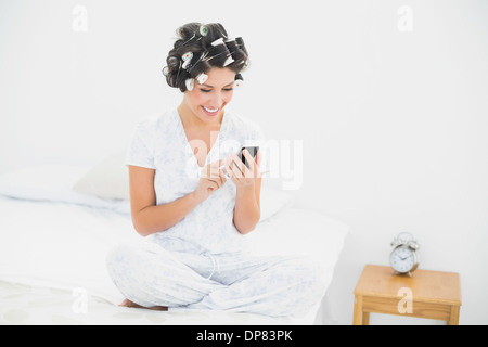 Cheerful brunette in hair rollers sending a text on bed Banque D'Images