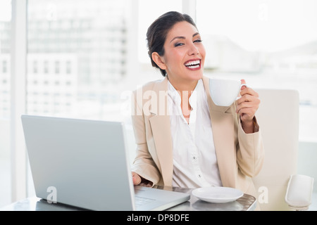 Laughing businesswoman holding cup tout en travaillant sur l'ordinateur portable Banque D'Images