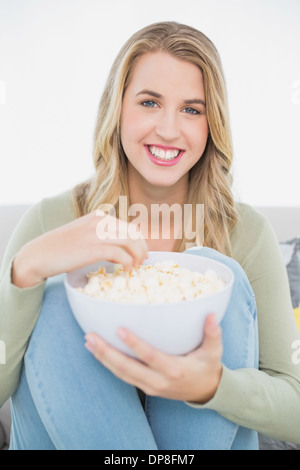 Cheerful pretty blonde eating popcorn sitting on cosy sofa Banque D'Images