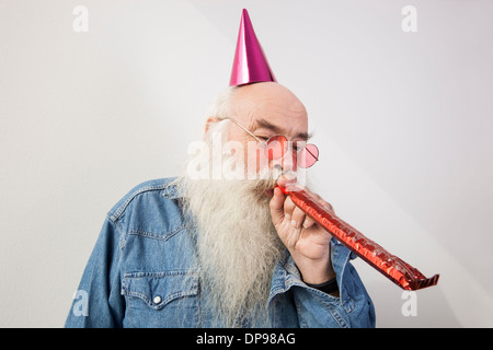 Senior man wearing party hat tout en soufflant corne contre fond gris Banque D'Images