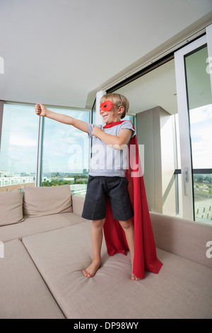 Smiling boy en costume de super-héros avec bras tendus permanent sur un canapé-lit à la maison Banque D'Images