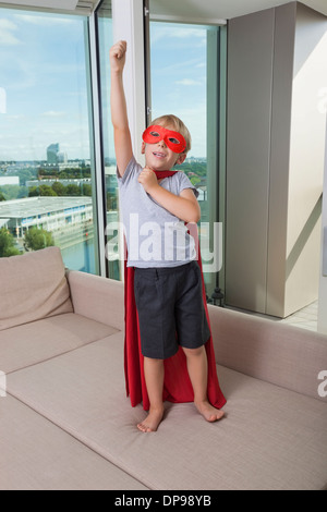 Garçon en costume de super héros debout avec bras levé sur un canapé-lit à la maison Banque D'Images