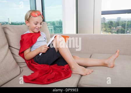 Smiling boy en costume de super-héro reading book on sofa at home Banque D'Images