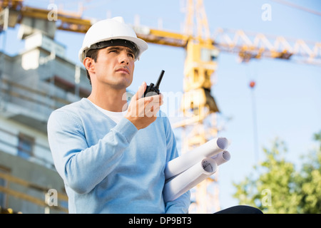 Male architect with blueprints à l'aide de talkie-walkie at construction site Banque D'Images