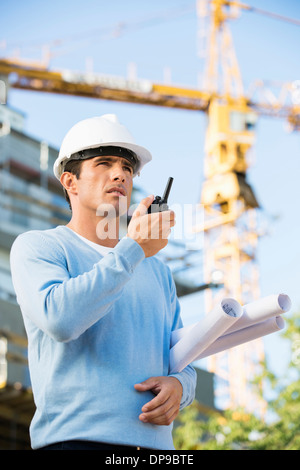 Male architect holding blueprints lors de l'utilisation de talkie-walkie at construction site Banque D'Images