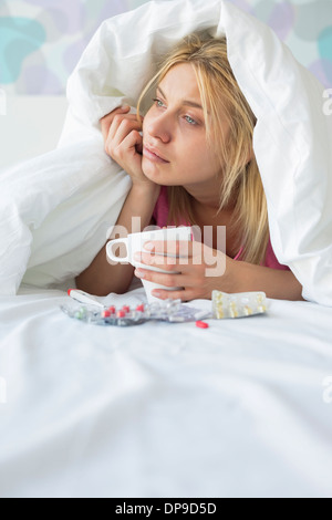 Young woman with coffee mug et médicaments souffrant de fièvre tandis que l'objet de couette sur lit Banque D'Images