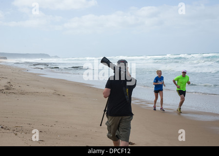 Jeune couple jogging sur la rive de la plage un jour d'été et un photographe portant un trépied en passant devant eux Banque D'Images