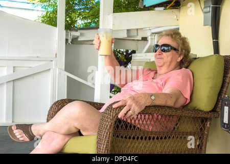 Une personne âgée, Caucasian woman est titulaire d'une île rum cocktail tout en étant assis sur une véranda sur une jolie plage, à Sainte-Croix, îles Vierges américaines. Banque D'Images